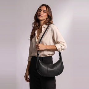 Femme portant le sac demi-lune en cuir Eugénie noir à l'épaule, à hauteur de hanche
